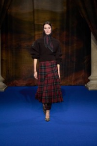 Mel Van Roemburg portant un pull en maille côtelée coloris brun Bark, un pantalon kilt plissé en rouge Poppy, des escarpins Cameo en coton brun Brush, ainsi qu'un bracelet Charm plaqué or et un foulard en soie à carreaux rouge Wine. 