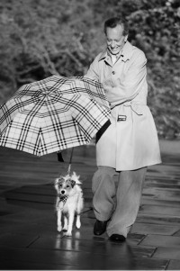 Richard Grant in einem Burberry-Trenchcoat und mit einem Taschenschirm in Check