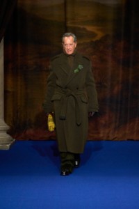 Richard E. Grant usando trench coat em lã e sarja Cavalry nas cores marrom Heath e verde Dutchy, suéter de lã canelado em marrom mesclado, camisa estilo pijama de seda em bege Clam, calças de veludo cotelê na cor verde meia-noite e botas de couro pretas. 