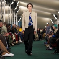 O modelo está usando uma jaqueta biker de couro, blusa xadrez em ponto canelado e calças de alfaiataria.