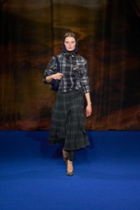 Saunders wearing mohair wool sweater in black and white, pleated check wool trousers in grey, check cotton pumps in brush brown, leather bag in midnight blue, Gold-plated Star Horse brooch, Silver Shield Segment ring and cashmere silk scarf in ink blue.