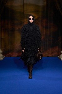 Hejia Li portant une cape noire à franges en maille crochetée de mohair et laine mélangés, un body noir en maille gaufrée de laine d'alpaga mélangée et une jupe noire plissée en soie à motif tournesol, avec des bottes Cavalier en cuir marron Truffle et un sac en cuir bleu Ink.