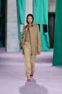 Modelo que luce chaqueta estilo trench coat color caqui y Milk con cremallera, camiseta sin mangas en tejido jersey de nailon color Hemlock y pantalones en lino color caqui y Milk.