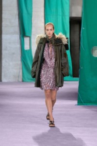 Model in Parka aus gewachster Baumwolle in der Farbe Oil und Jerseykleid aus Viskose in der Farbe Lilac mit Fransen und Pailletten in Metallic-Optik