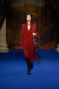 Chu Wong portant une robe en damas de velours baroque rouge Dragon, des bottes Cavalier en cuir noir, un sac Highlands en brun Hazel, ainsi qu'un bracelet et un collier Charm plaqué or.