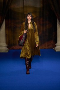 Neelam Gill usando vestido de veludo em jacquard com estampa geométrica em amarelo pera, botas de cano alto de comprimento até os quadris em vermelho mogno, bolsa de ombro Highlands em couro na cor roxo figo, pulseira Knight com pingente folheada a ouro e anel Hollow.