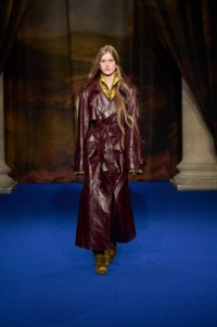 Iris Lasnet wearing ornate embossed leather trench coat in brown, pyjama shirt and trousers in pear yellow and Cameo slippers in cub brown and Star earrings.