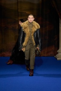 Sang Woo Kim vestindo jaqueta aviador de shearling preta, paletó e calças moulinè nas cores cinza Tarmac e bege Pebble, suéter de gola rulê em marrom cáqui e botas de couro em vermelho mogno.