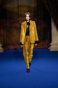 Dana Smith portant une veste et un pantalon en velours baroque coloris jaune Pear, un haut en velours noir et des escarpins Cameo en cuir embossé à franges et à motif ornemental de couleur rouge Mahogany.