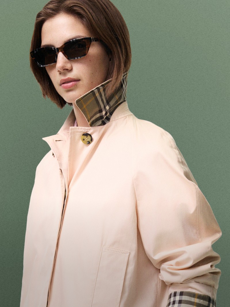 Person wearing oversized sunglasses and a light pink jacket with a check-pattern collar against a muted green background.