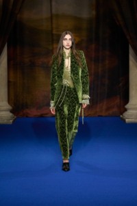 Amelie Steele portant une veste et un pantalon tailleur en damas de velours baroque vert, avec des escarpins Cameo à franges en cuir embossé de couleur vert Moat.