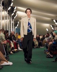 O modelo está usando uma jaqueta biker de couro, blusa xadrez em ponto canelado e calças de alfaiataria.