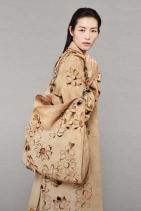 Model in Trenchcoat und Hose aus Veloursleder mit Gänseblümchen-Cutout-Details, gestylt mit kleiner Henkeltasche „Shield Twin“ mit Gänseblümchen-Cutout-Details